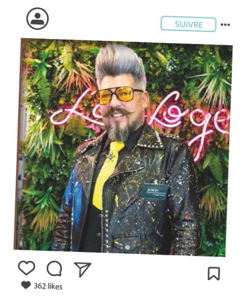 Portait de Joris Davoine, portant une chevelure Gris argentée, coiffé en hauteur, ab=vec une barbe et moustache de la même couleur. Il porte une seste en cuir et des lunettes aux verres teintés jaune, look rock avec son perfecto